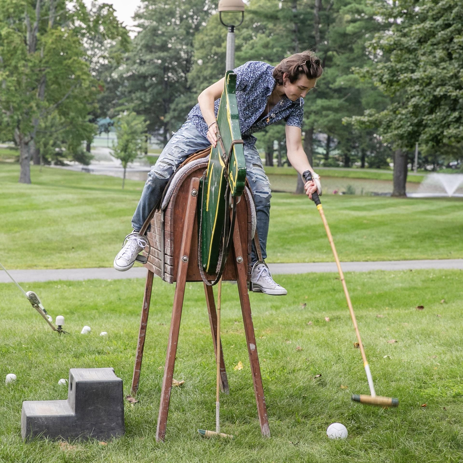A student sits on a fake horse and reaches down to hit a polo ball with a mallet. 